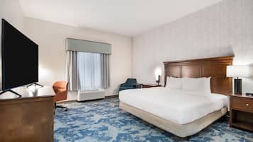 A hotel room with a large bed, a desk with a flat-screen TV, a chair, a window with curtains, and a wall-mounted lamp.