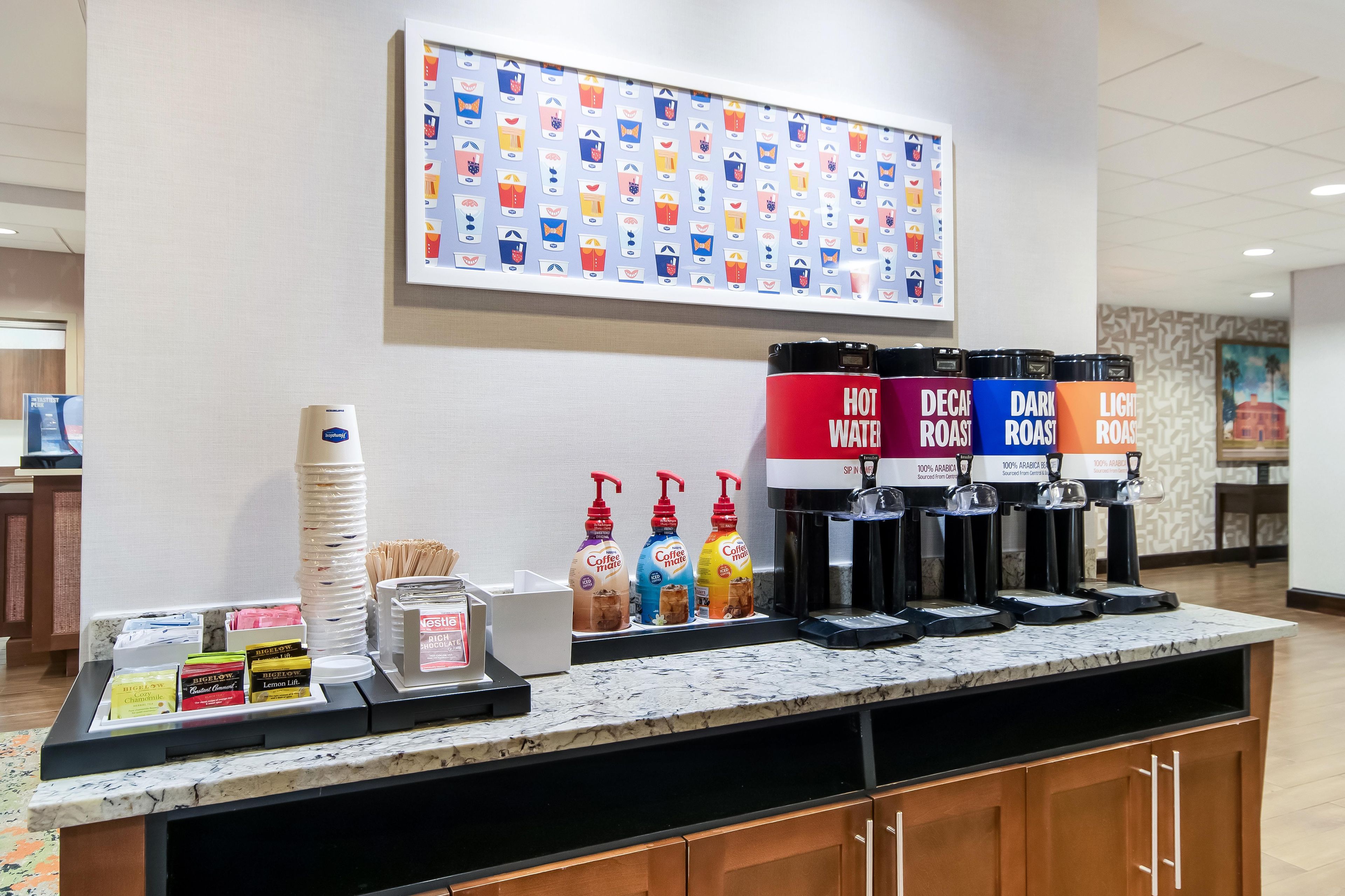 A coffee station with multiple dispensers, a coffee menu, and a wall with beverage icons.