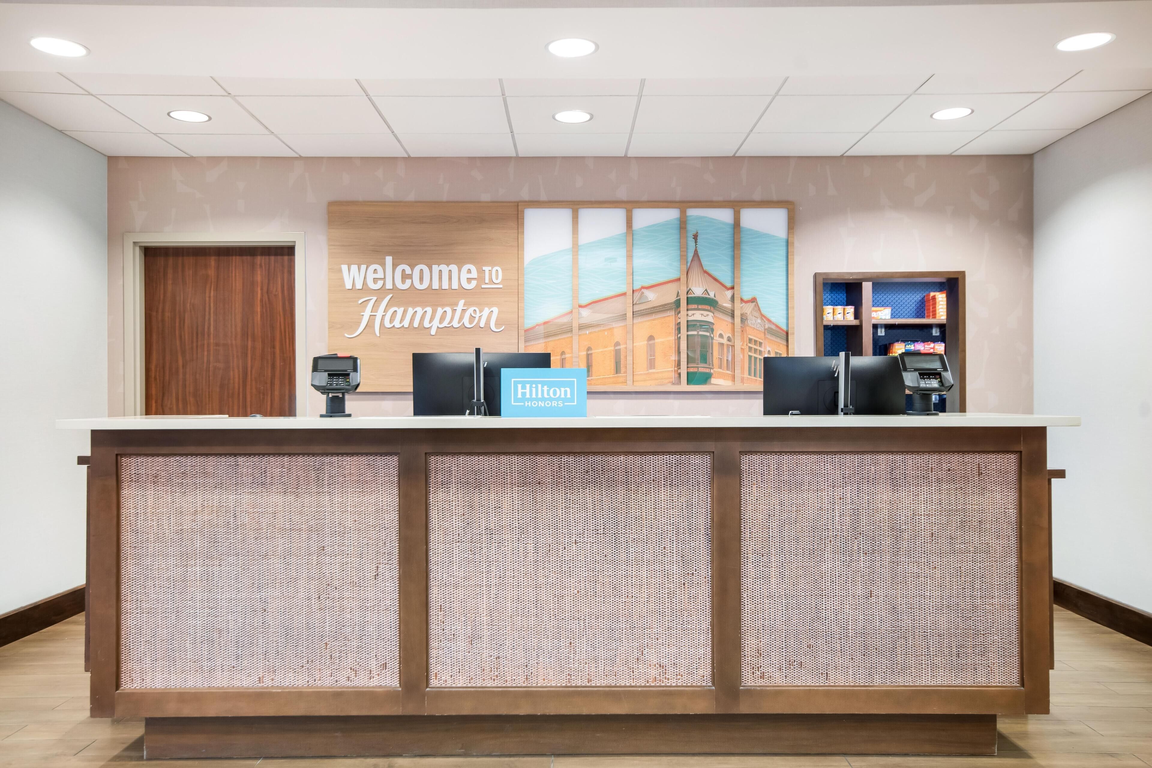 A reception desk with a 'Welcome to Hampton' sign, a Hilton Honors badge, a phone, and a small shelf with items.