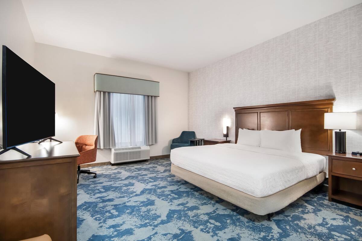 A hotel room with a large bed, a desk with a flat-screen TV, a chair, a window with curtains, and a wall-mounted lamp.