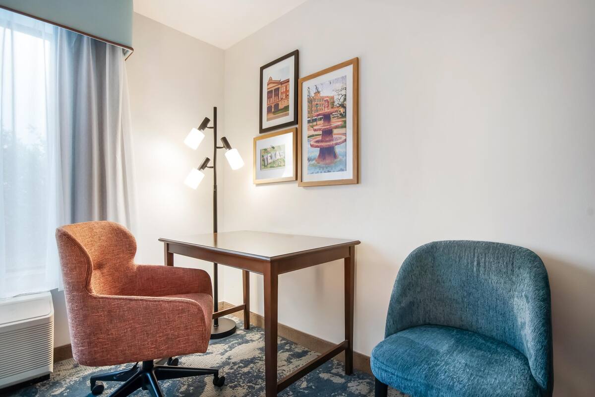 A hotel room with a desk, two chairs, a standing lamp, and three framed pictures on the wall.