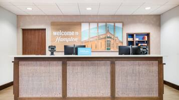 A reception desk with a 'Welcome to Hampton' sign, a Hilton Honors badge, a phone, and a small shelf with items.