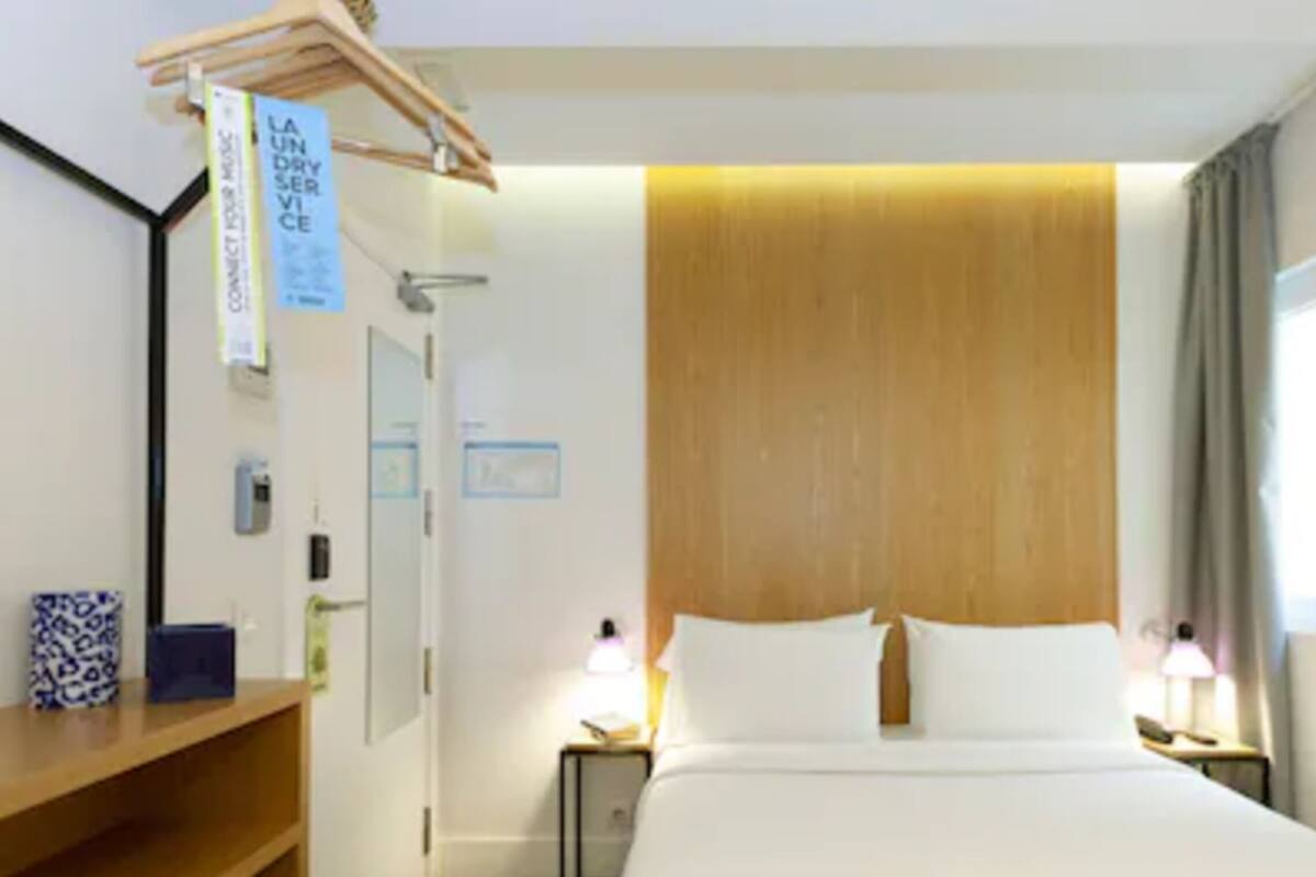 A hotel room with a wooden headboard, a bed with white linens, a nightstand with a lamp, a wooden shelf with a decorative item, and a window with curtains.