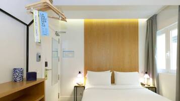 Habitación de hotel con cabecera de madera, cama con ropa blanca, mesita de noche con lámpara, estante de madera con un objeto decorativo y ventana con cortinas.