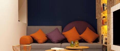 Una sala moderna con un sofá gris, cojines naranja y morados, una mesa de centro de madera y una mesa de centro de vidrio. Hay una estantería con libros y objetos decorativos.