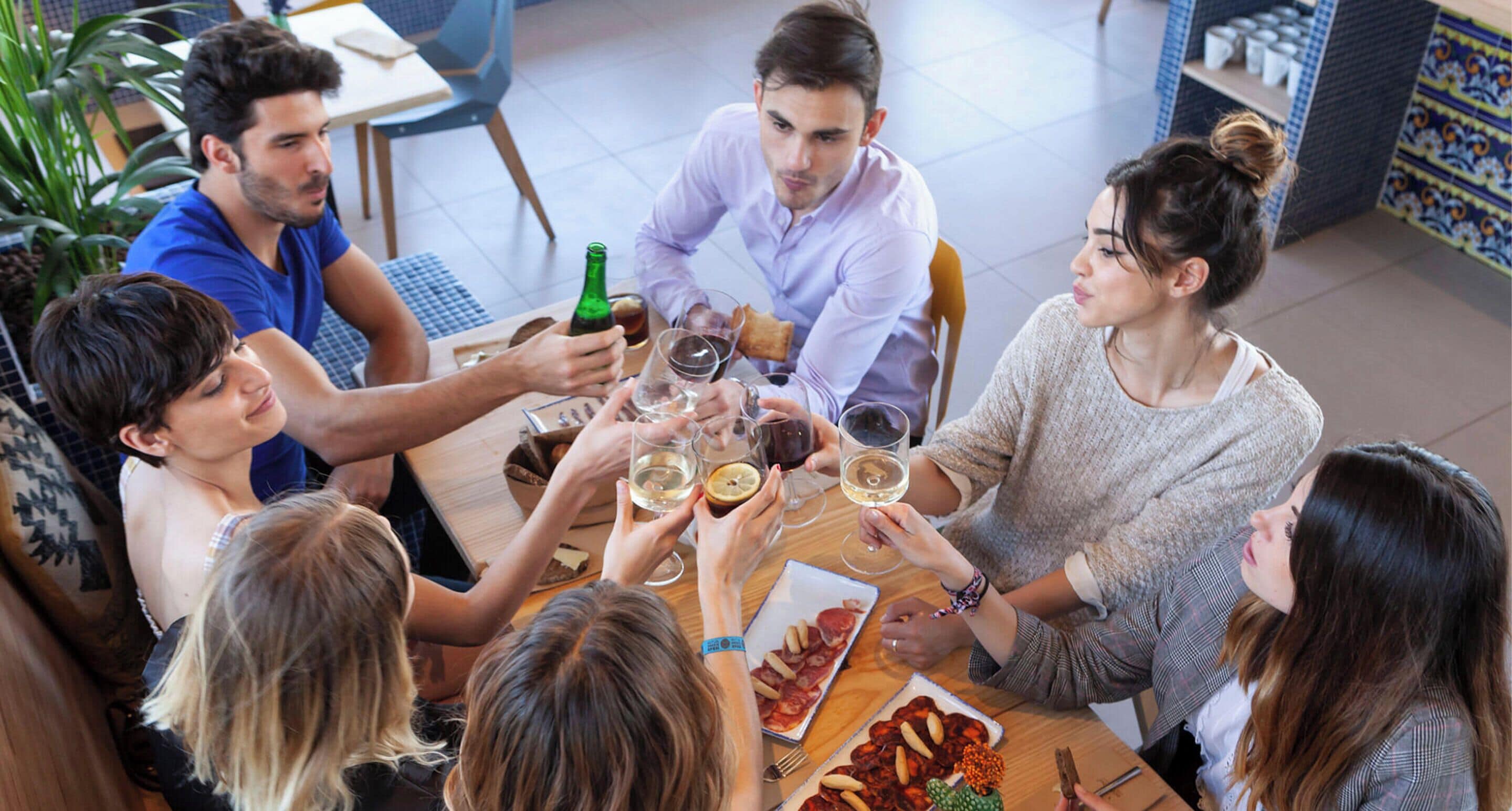 A group of people sitting around a table with food and drinks, in a room with tiled flooring and a plant in the background.
