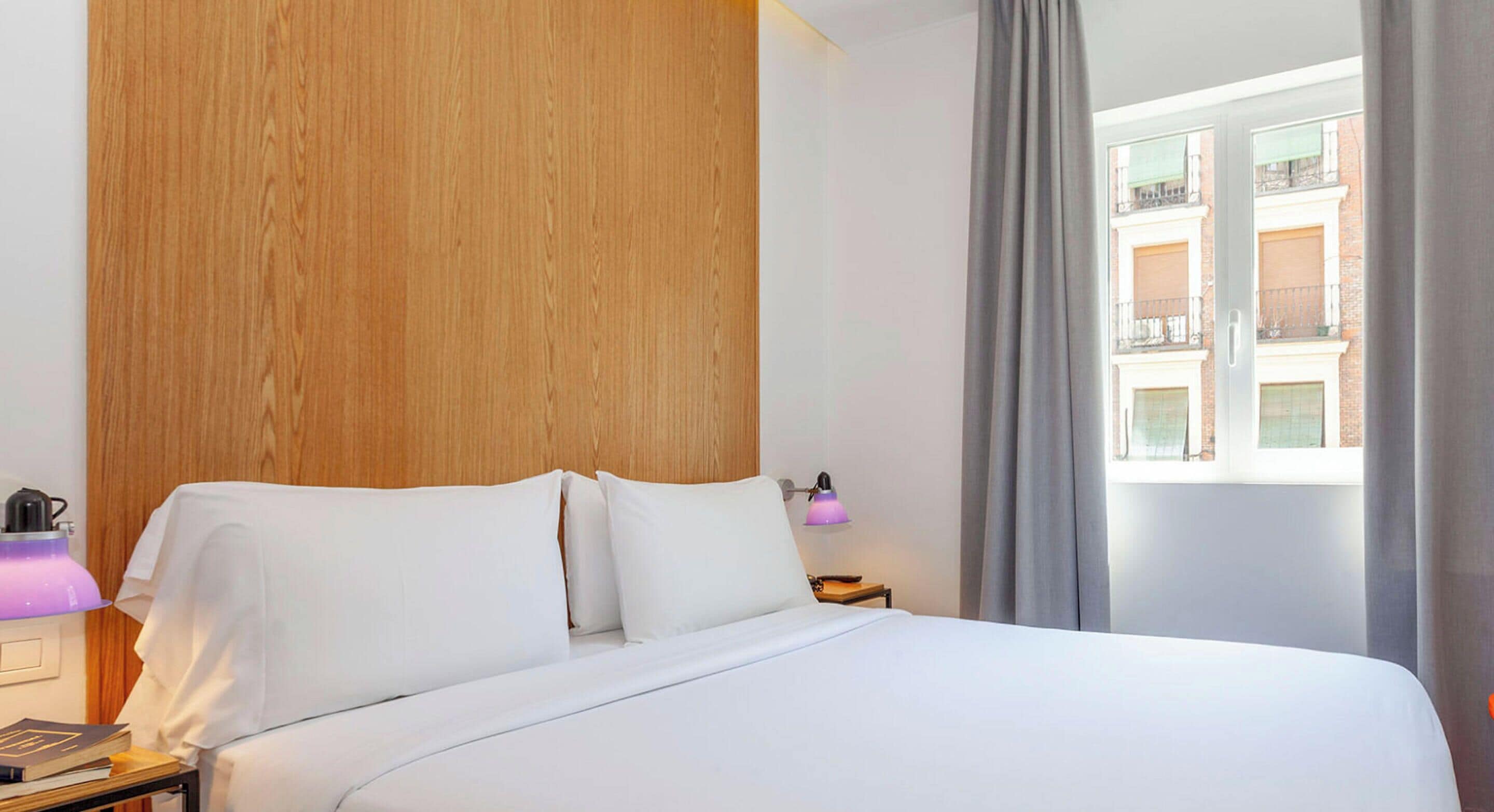 Habitación de hotel con cabecera de madera, cama con ropa de cama blanca, mesita de noche con lámpara y ventana con vistas a edificios.