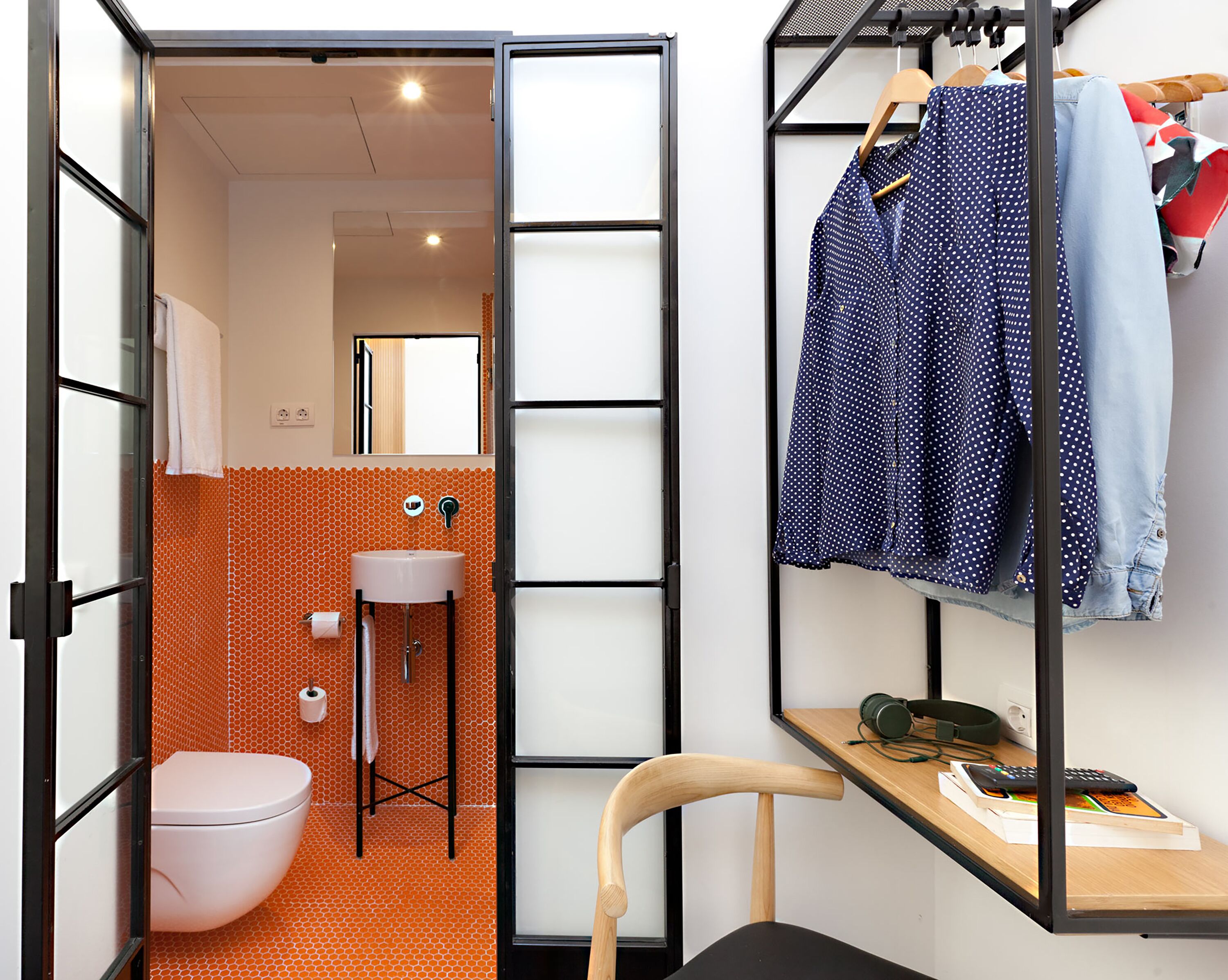 A compact, modern bathroom with a toilet, sink, and shower. There is a wooden chair and a small table with a phone and remote control. Clothes are hanging on a rack.