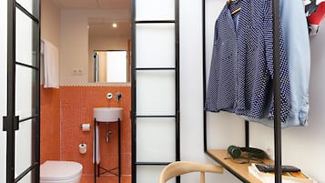 Baño moderno y compacto con inodoro, lavamanos y ducha. Hay una silla de madera y una mesita con un teléfono y un control remoto. Ropa colgada en un perchero.