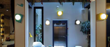 A modern hotel lobby with a central elevator, seating area, and decorative lighting.