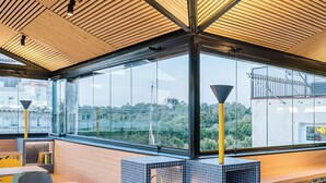 A modern restaurant with a high ceiling, large windows, and a view of the city. It features a bar area with stools and a seating area with tables and chairs.