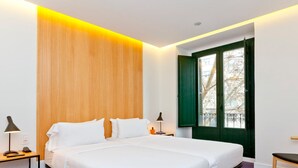 A modern hotel room with a large bed, bedside tables with lamps, a window with green shutters, and a wooden accent wall.