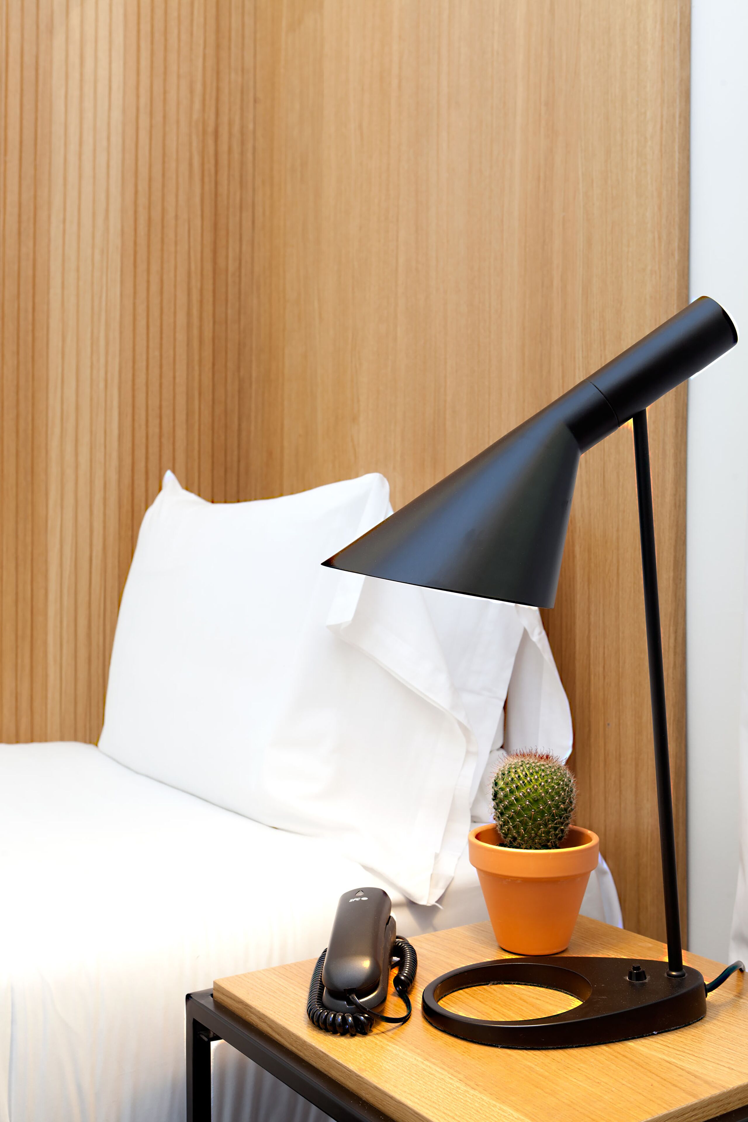 A neatly made bed with white bedding, a black telephone, a black desk lamp, a small potted cactus, and a wooden nightstand.