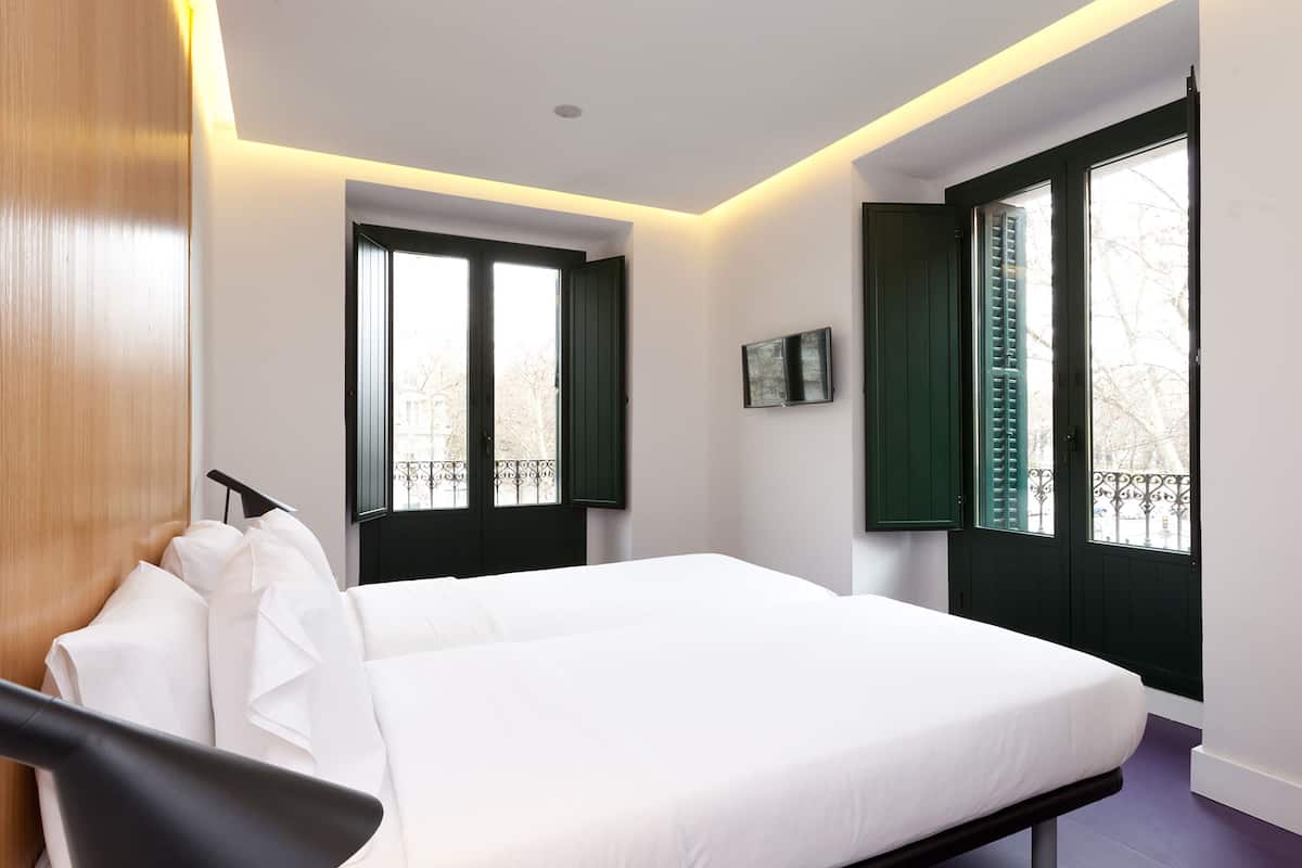 A modern bedroom with a large bed, two windows with shutters, a mounted TV, and a wooden door.