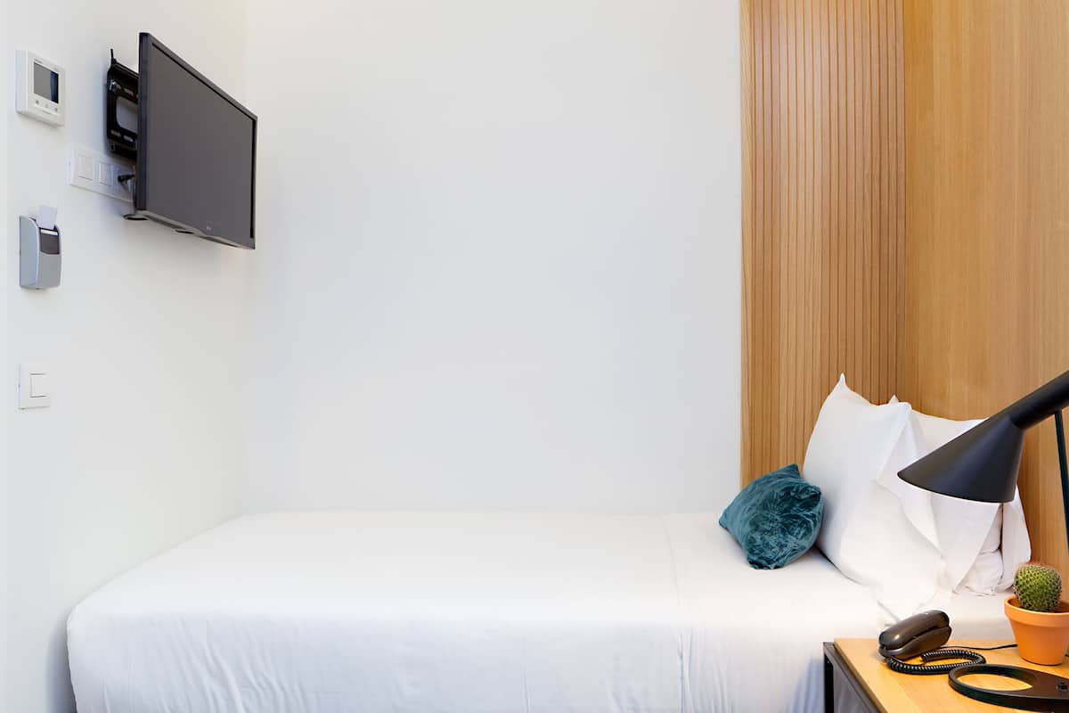 A neatly made bed with white linens, a mounted flat-screen TV, a bedside lamp, a small potted plant, and a telephone on a wooden nightstand.