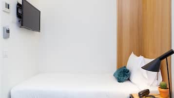 Una cama bien hecha con ropa blanca, un televisor de pantalla plana montado, una lámpara de noche, una pequeña planta en maceta y un teléfono sobre una mesita de noche de madera.