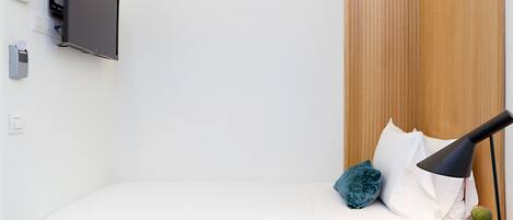 Una cama bien hecha con ropa blanca, un televisor de pantalla plana montado, una lámpara de noche, una pequeña planta en maceta y un teléfono sobre una mesita de noche de madera.