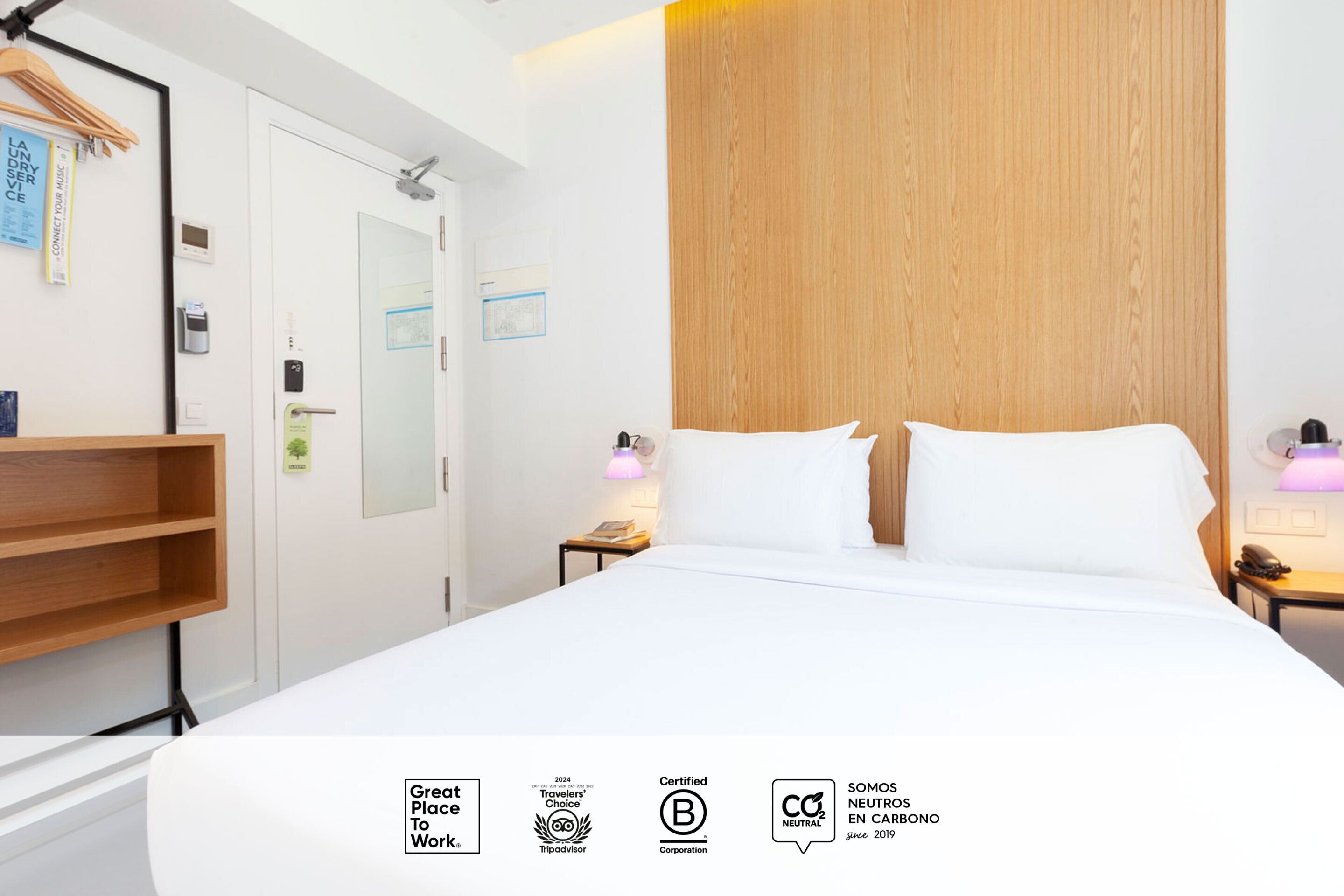 A hotel room with a bed, wooden headboard, bedside tables with lamps, a wooden shelf, and a door with a handle.