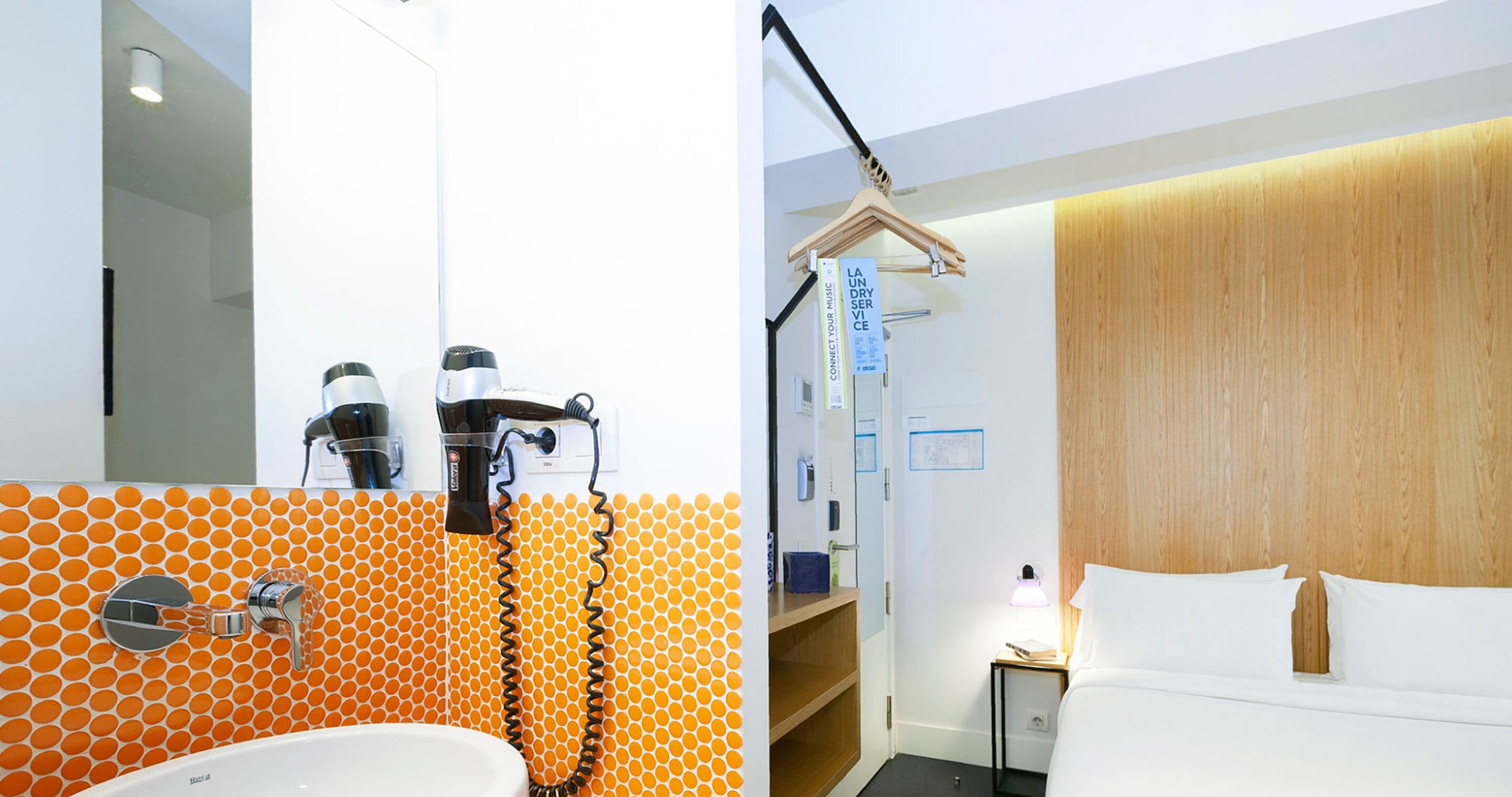 Baño con lavamanos, dos secadores de pelo y un espejo. La habitación tiene un revestimiento de madera y una cama con ropa blanca.
