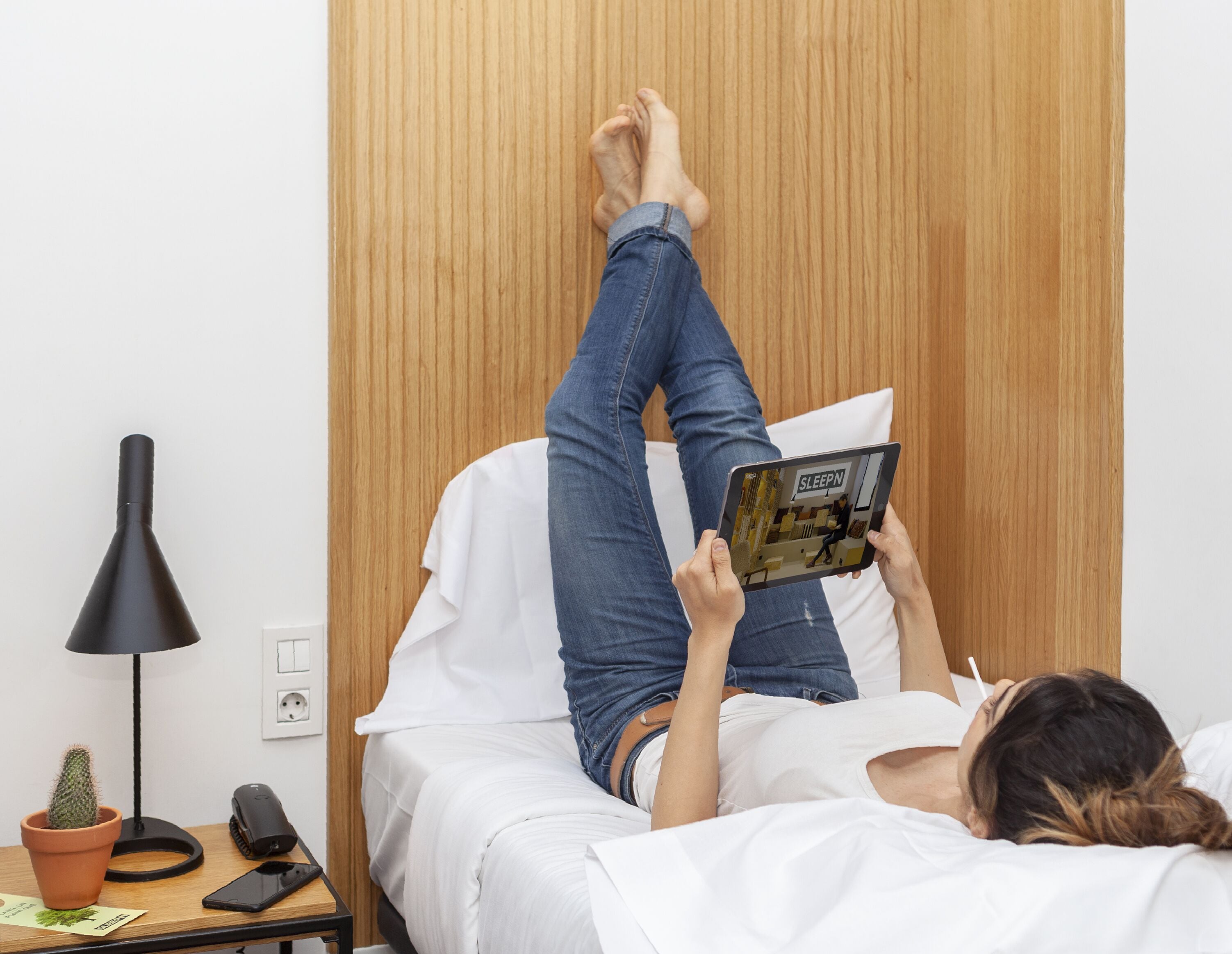 A person lying on a bed in a room with a wooden wardrobe, a desk with a lamp, a cactus, and a smartphone.