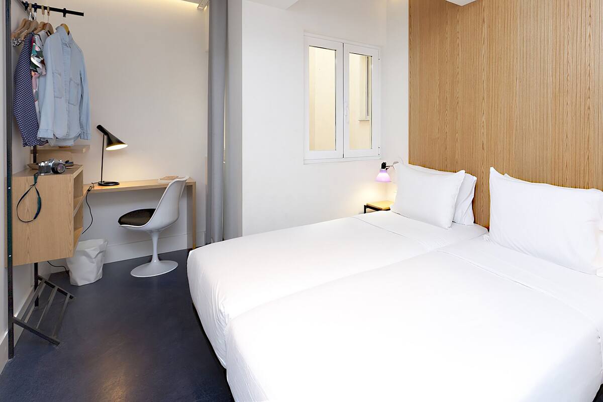 A hotel room with a bed, a desk with a chair, a wardrobe, and a window.