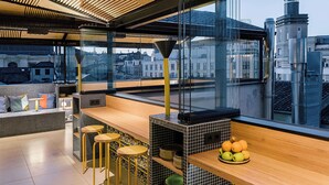 A modern bar area with a wooden counter, stools, and a view of the city through large windows.