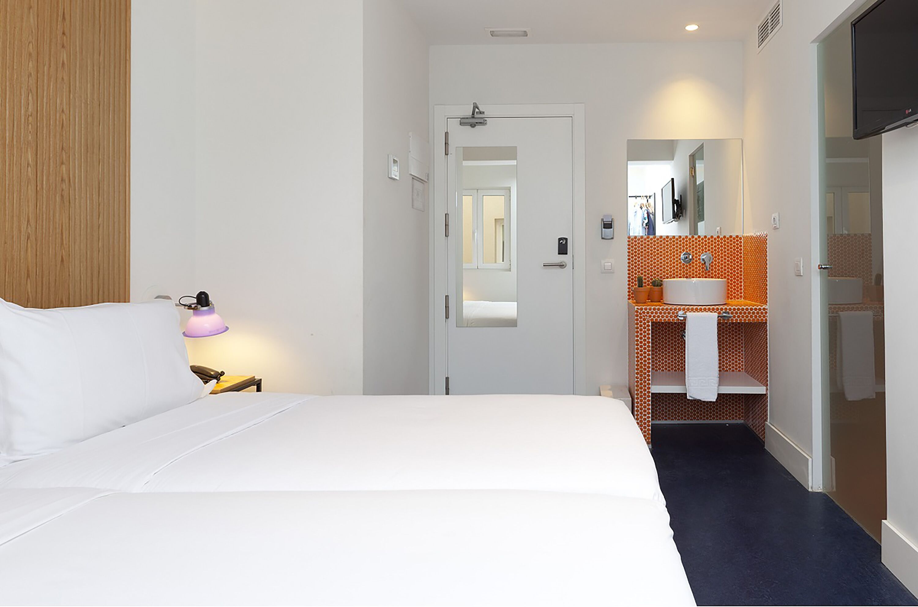 Habitación de hotel con cama, lámpara de noche, baño con lavabo y espejo, y puerta que da a otra habitación.