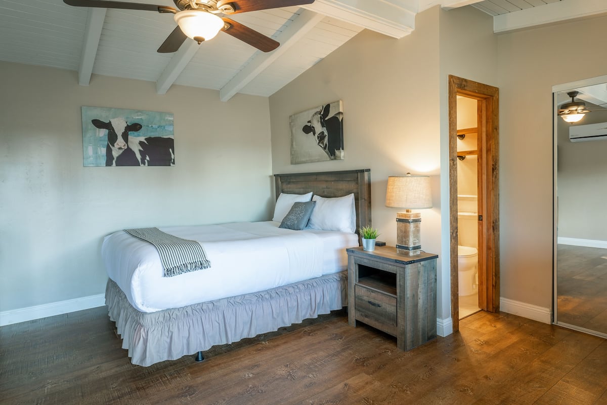 A bedroom with a large bed, wooden headboard, two wall-mounted paintings, a bedside table with a lamp, and a wooden floor.