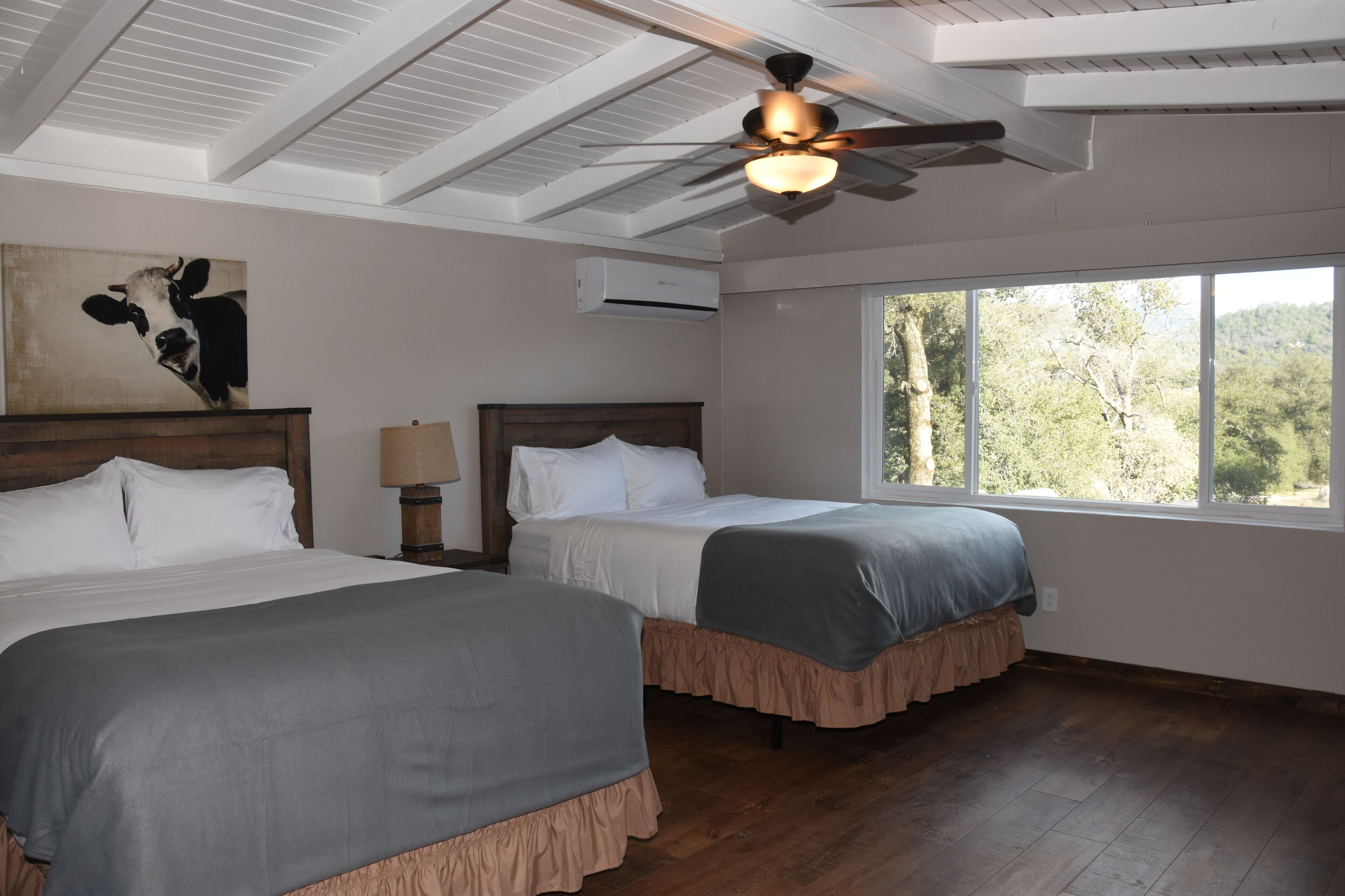 A room with two beds, a ceiling fan, a large window, and a cow painting on the wall.
