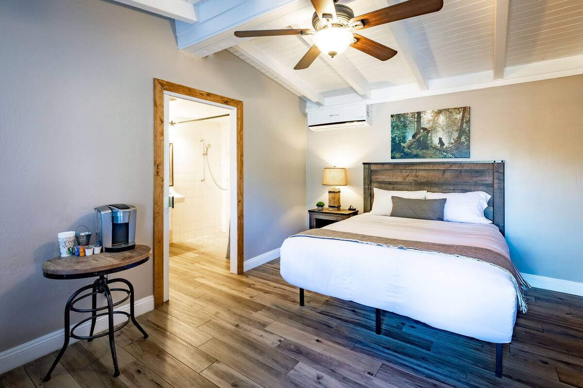 A bedroom with a wooden floor, a bed with white bedding, a ceiling fan, a nightstand with a lamp, and a painting on the wall.