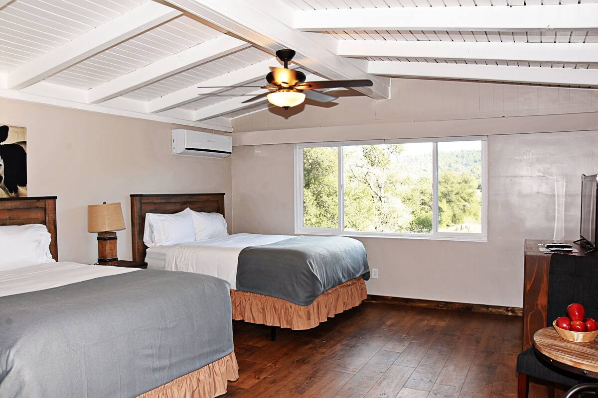 A hotel room with two beds, a ceiling fan, a large window with a view of trees, and a wooden floor.