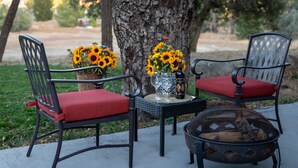 Two outdoor chairs with red cushions, a small table with a vase of sunflowers, and a fire pit.
