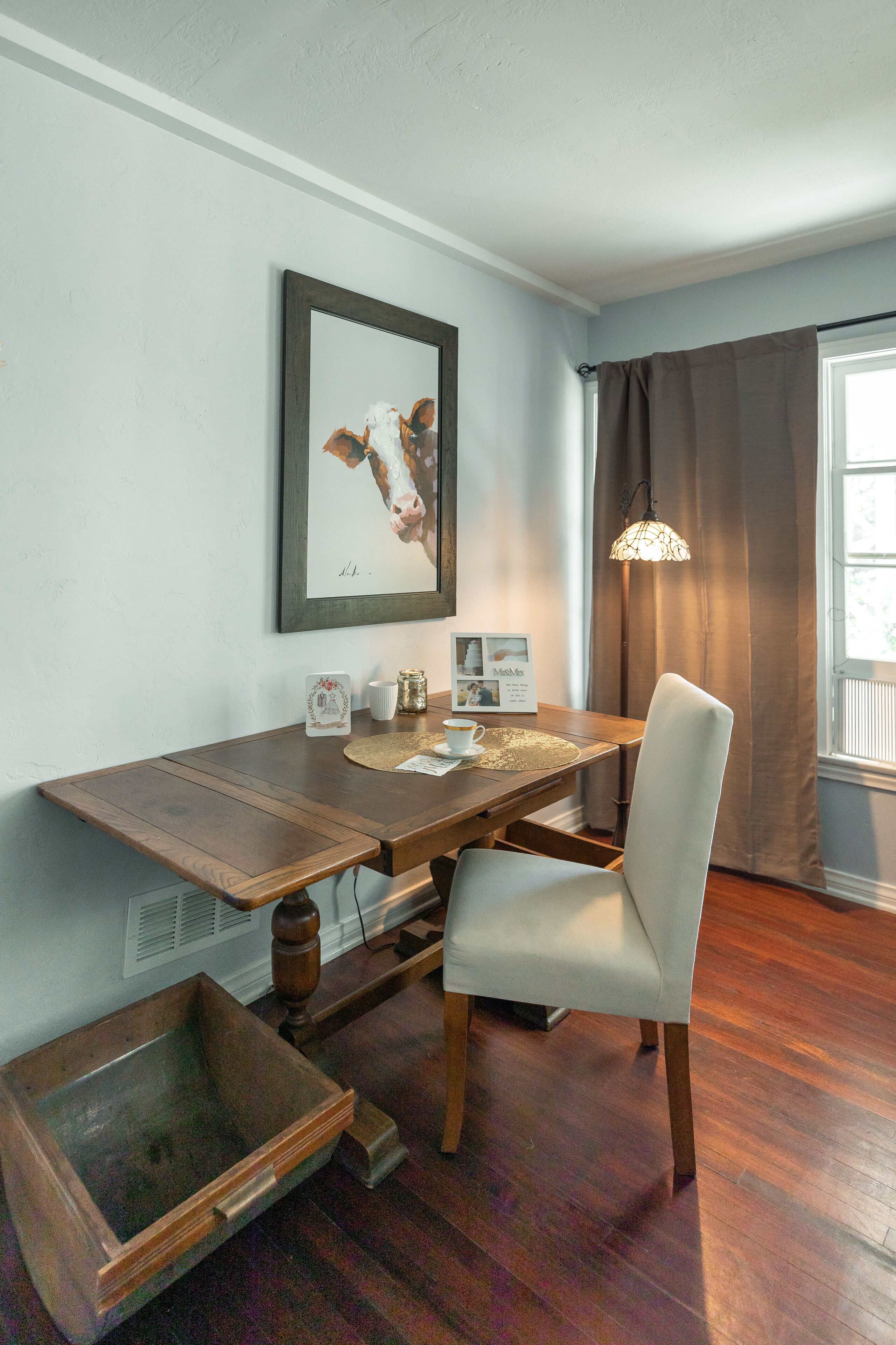 Un coin repas avec une table en bois, une chaise blanche, un tableau encadré, une bougie et une fenêtre avec des rideaux.
