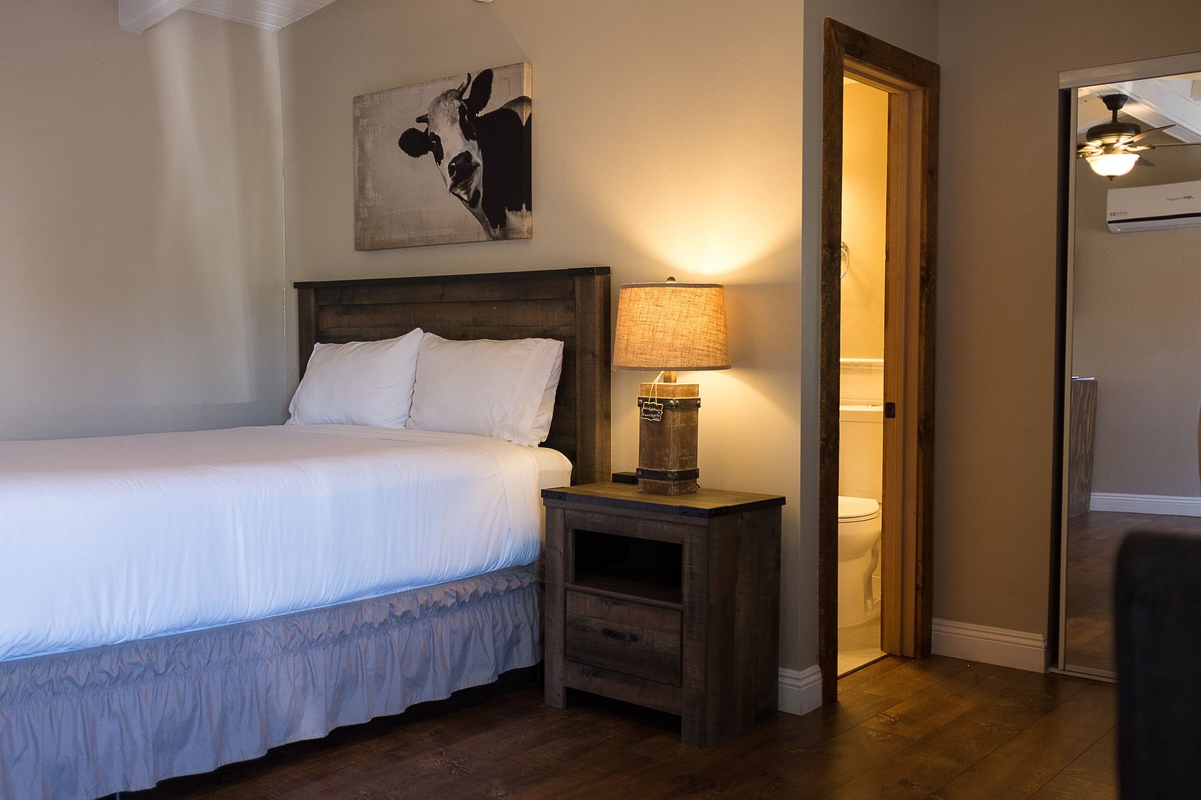 A bedroom with a bed, a nightstand with a lamp, a painting on the wall, and a door leading to a bathroom.