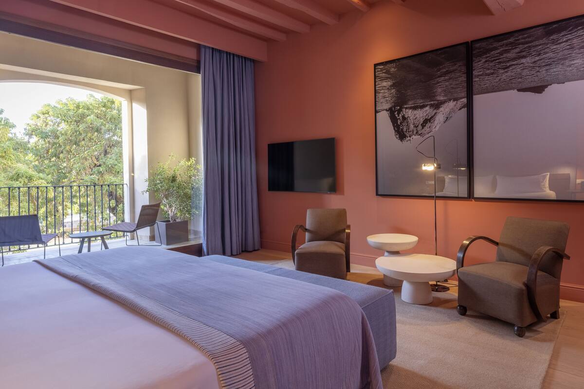 A modern hotel room with a large bed, two armchairs, a small round table, and a balcony with a view of trees.
