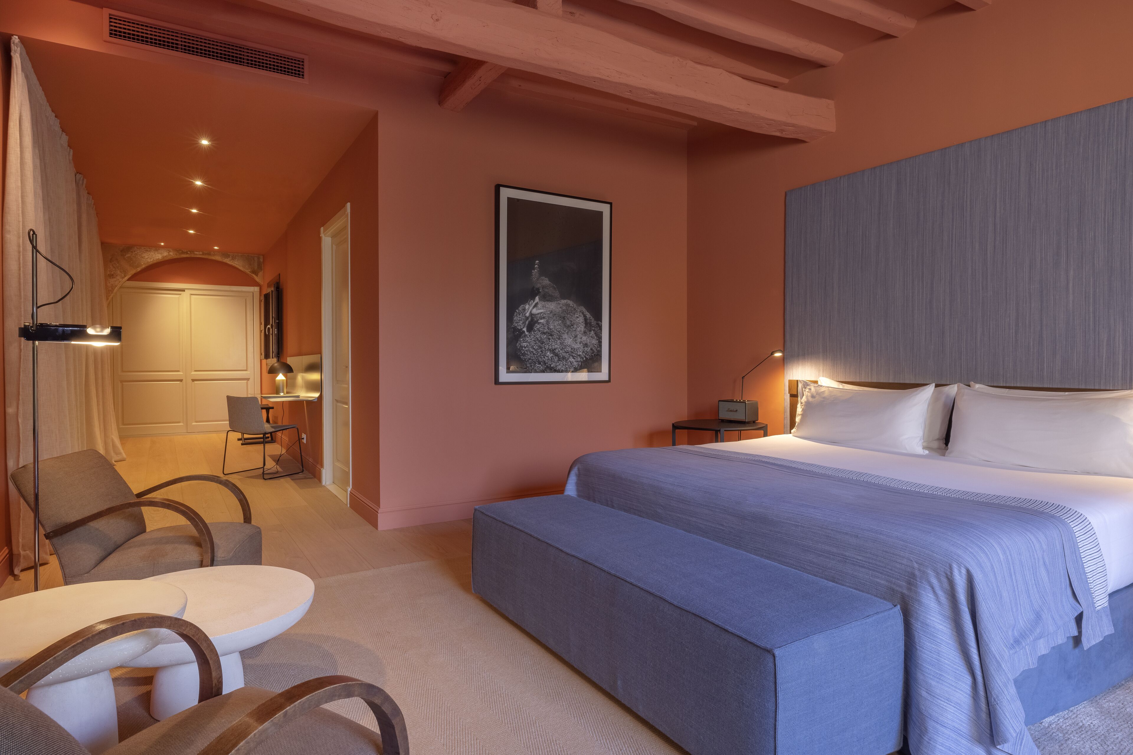 A bedroom with a large bed, a bedside table, a chair, and a desk with a lamp.