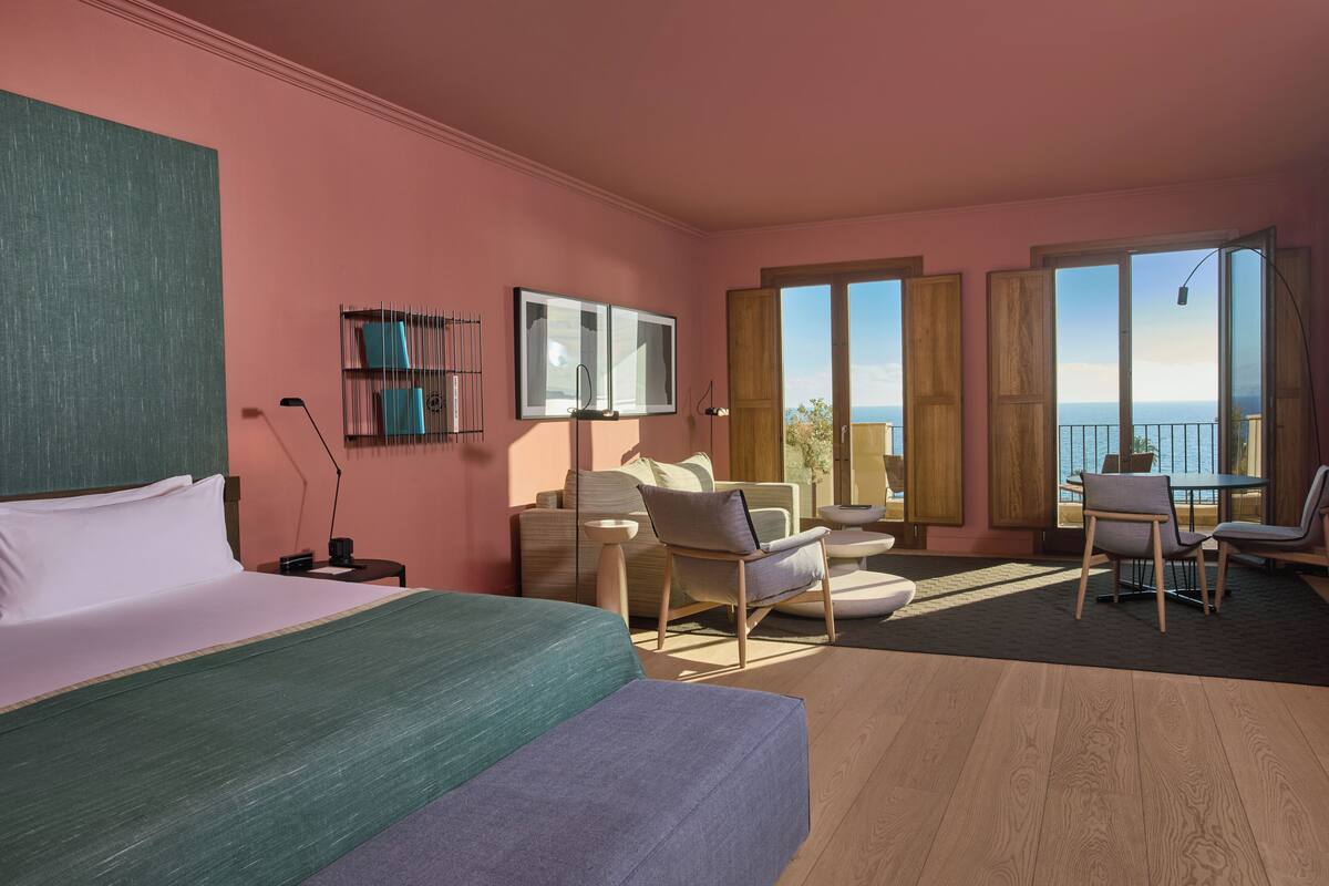 A modern hotel room with a large bed, a balcony with a view, and a seating area with a table and chairs.