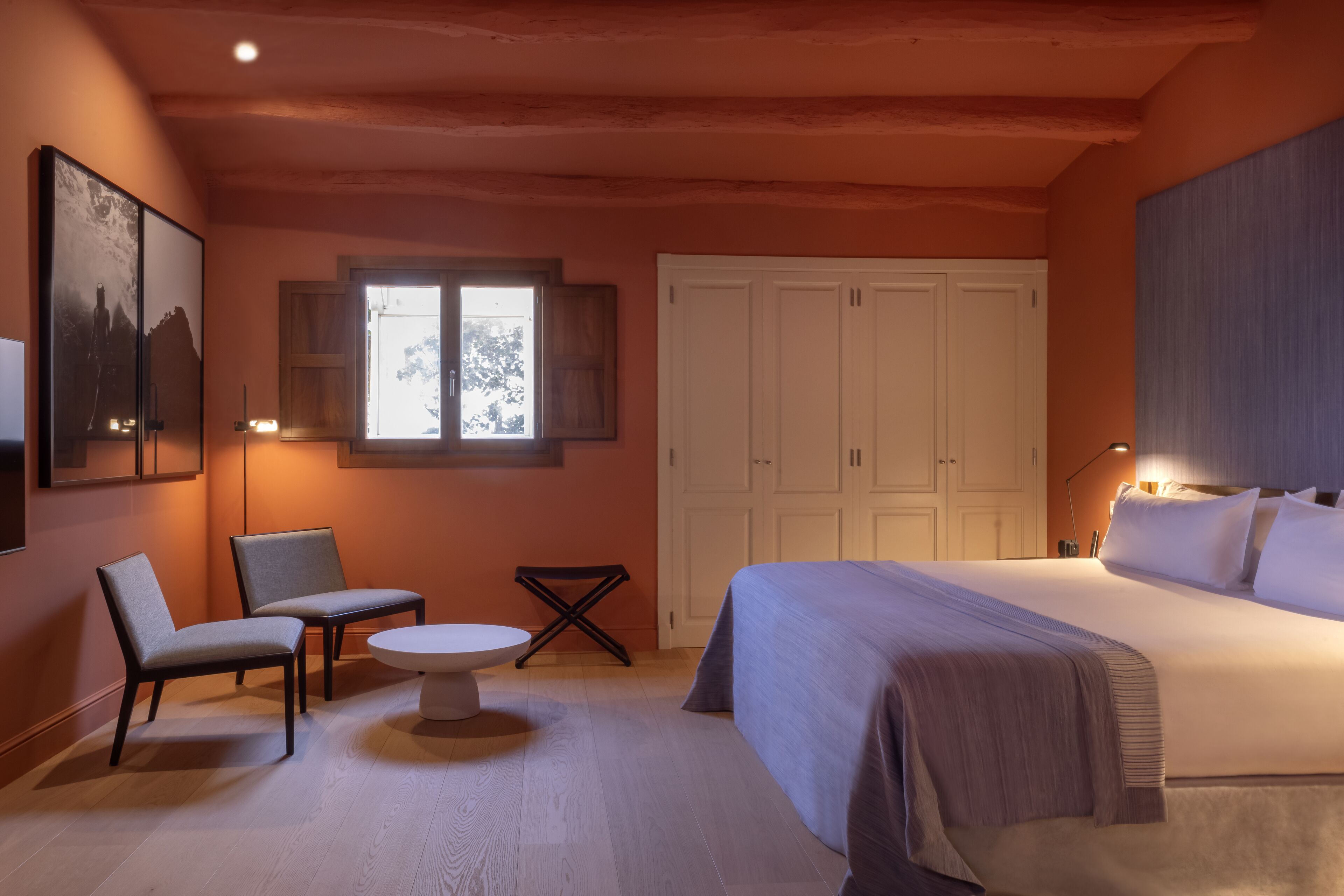 A bedroom with a bed, two chairs, a small table, a wardrobe, and a window.