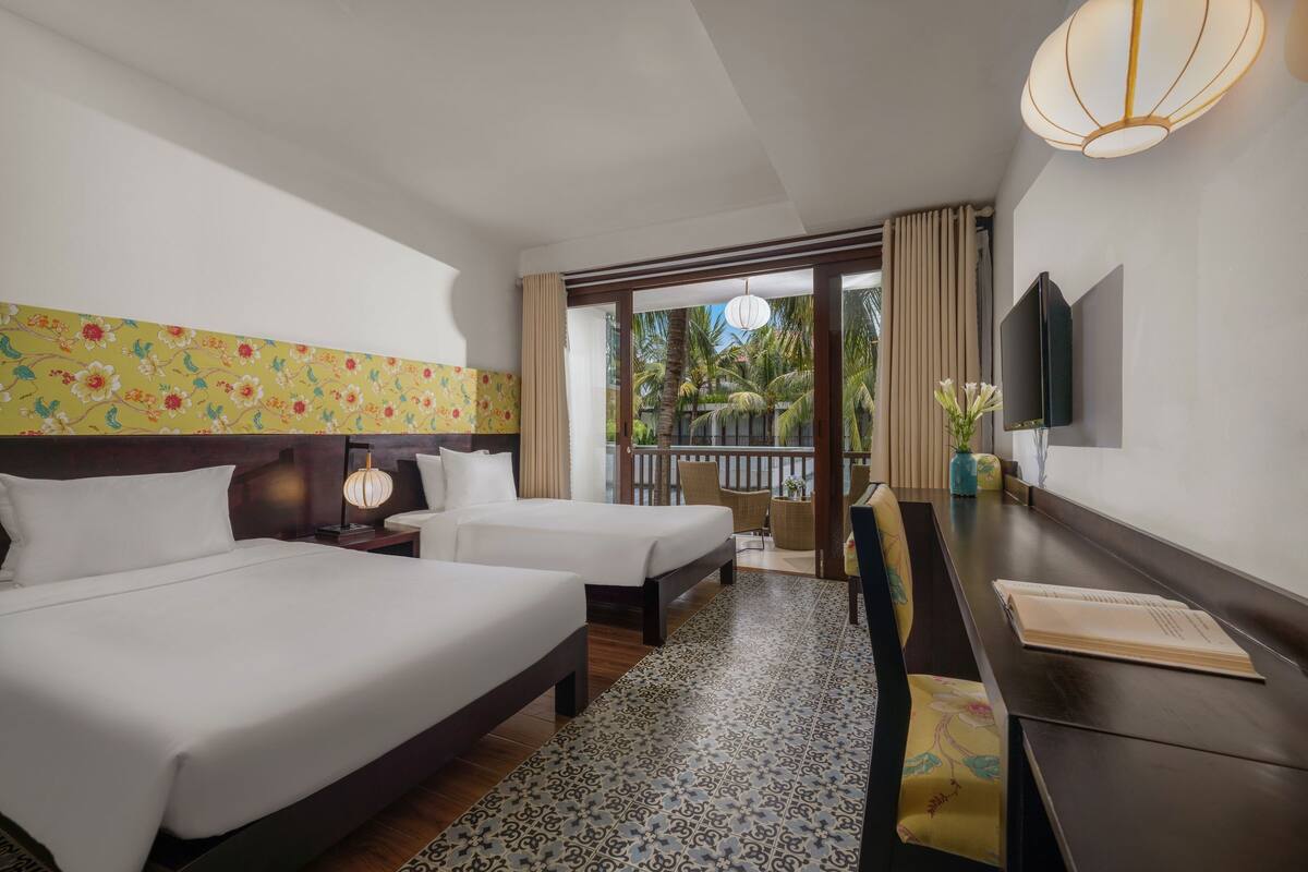 A hotel room with two beds, a desk, a TV, and a balcony with a view of palm trees.