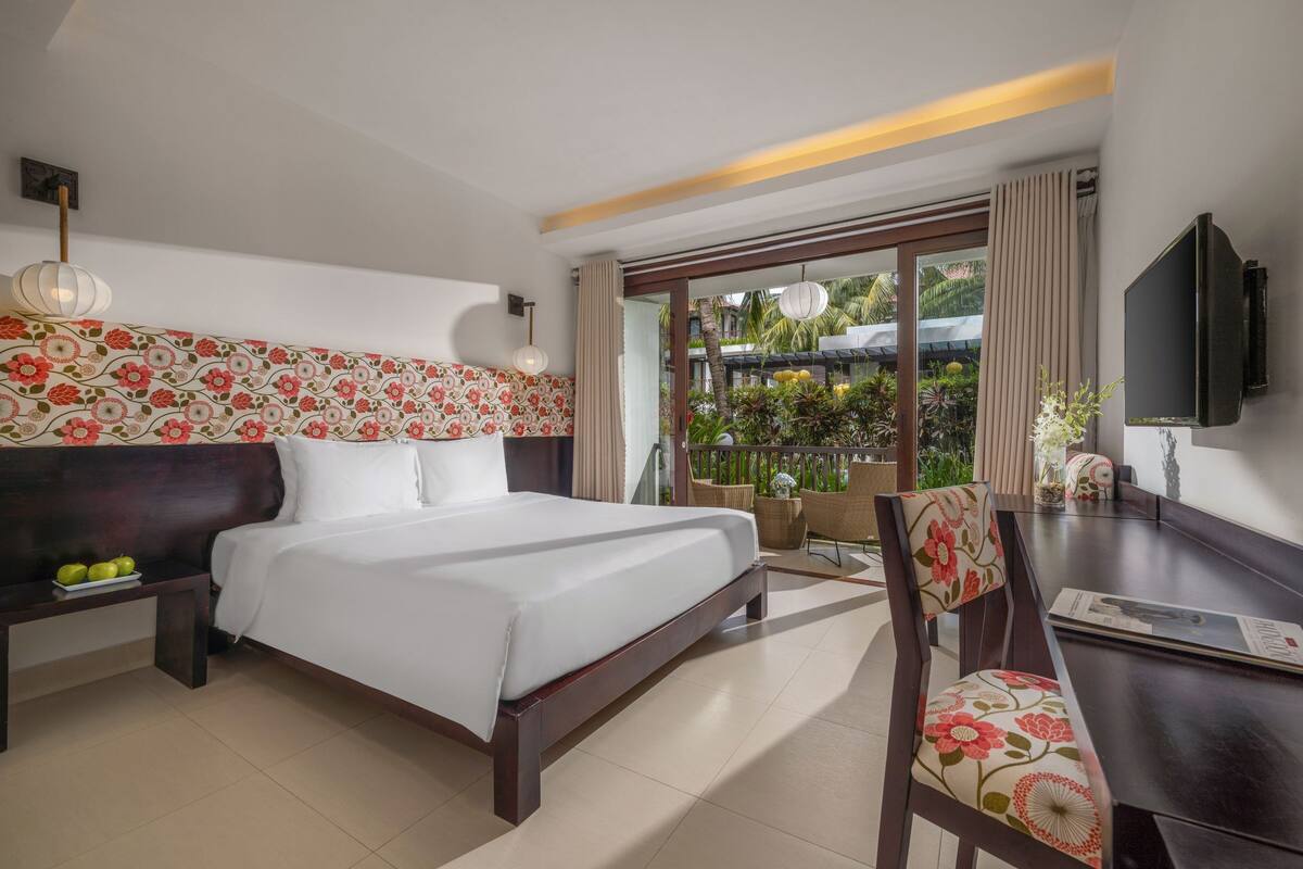A hotel room with a large bed, a desk with a chair, a TV, and a balcony with plants.