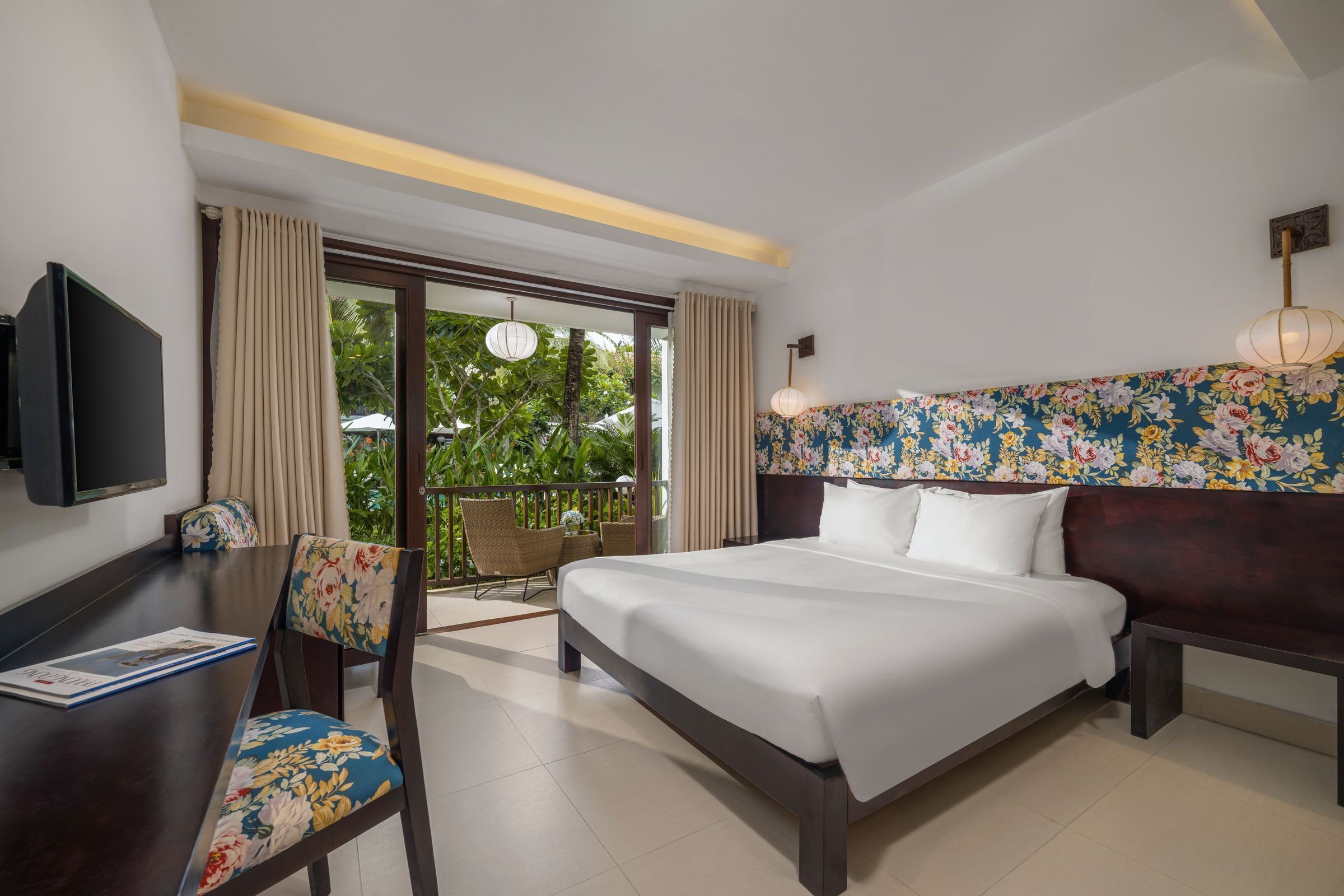 A hotel room with a large bed, a desk with a chair, a TV, and a balcony with a view of greenery.