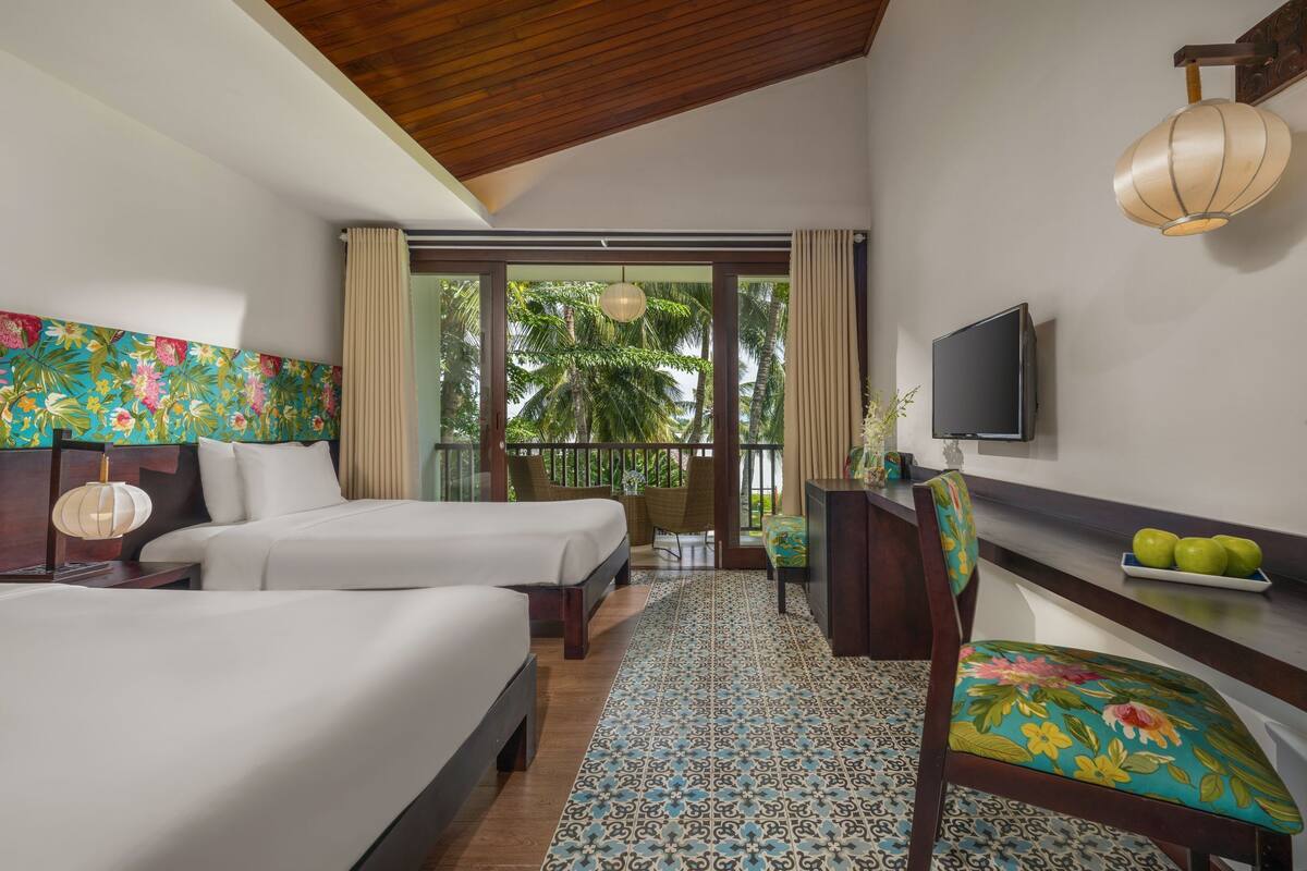 A hotel room with two beds, a TV, a dining area with a table and chairs, and a balcony with a view of palm trees.