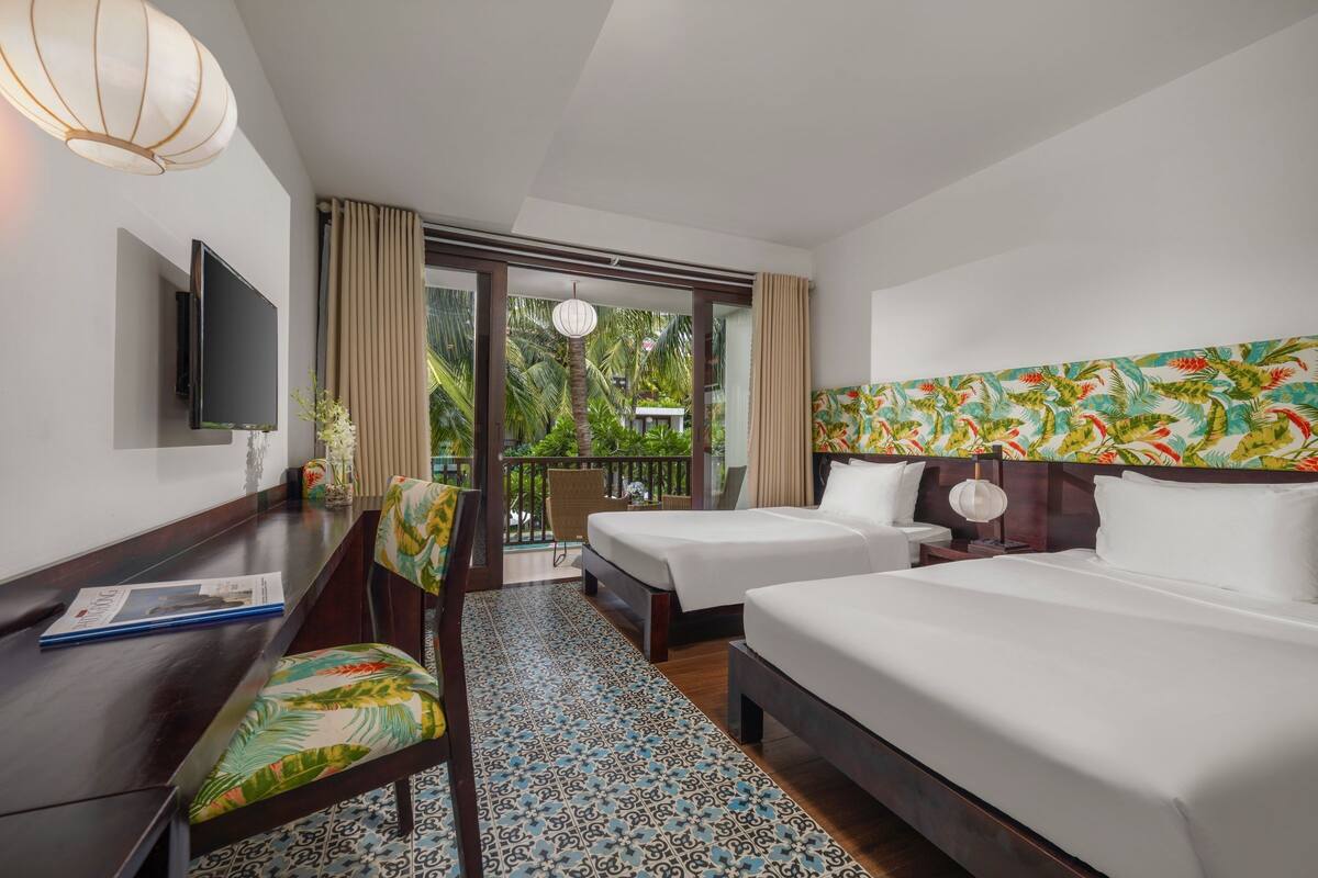 A hotel room with two beds, a desk, a TV, and a balcony with a view of greenery.
