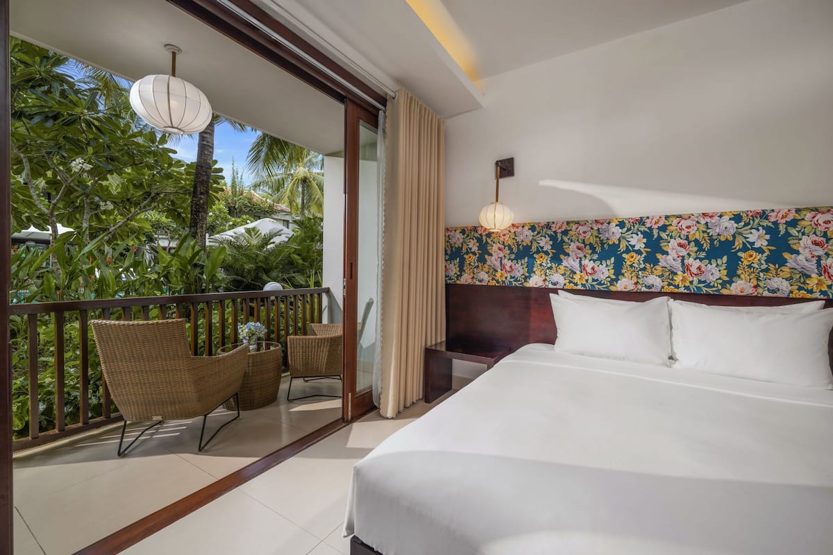 A hotel room with a balcony, a bed, wicker chairs, a small table, and a floral-patterned headboard.