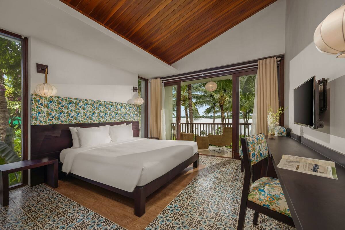 A hotel room with a large bed, a TV, a dining table with chairs, and a balcony with a view of palm trees.