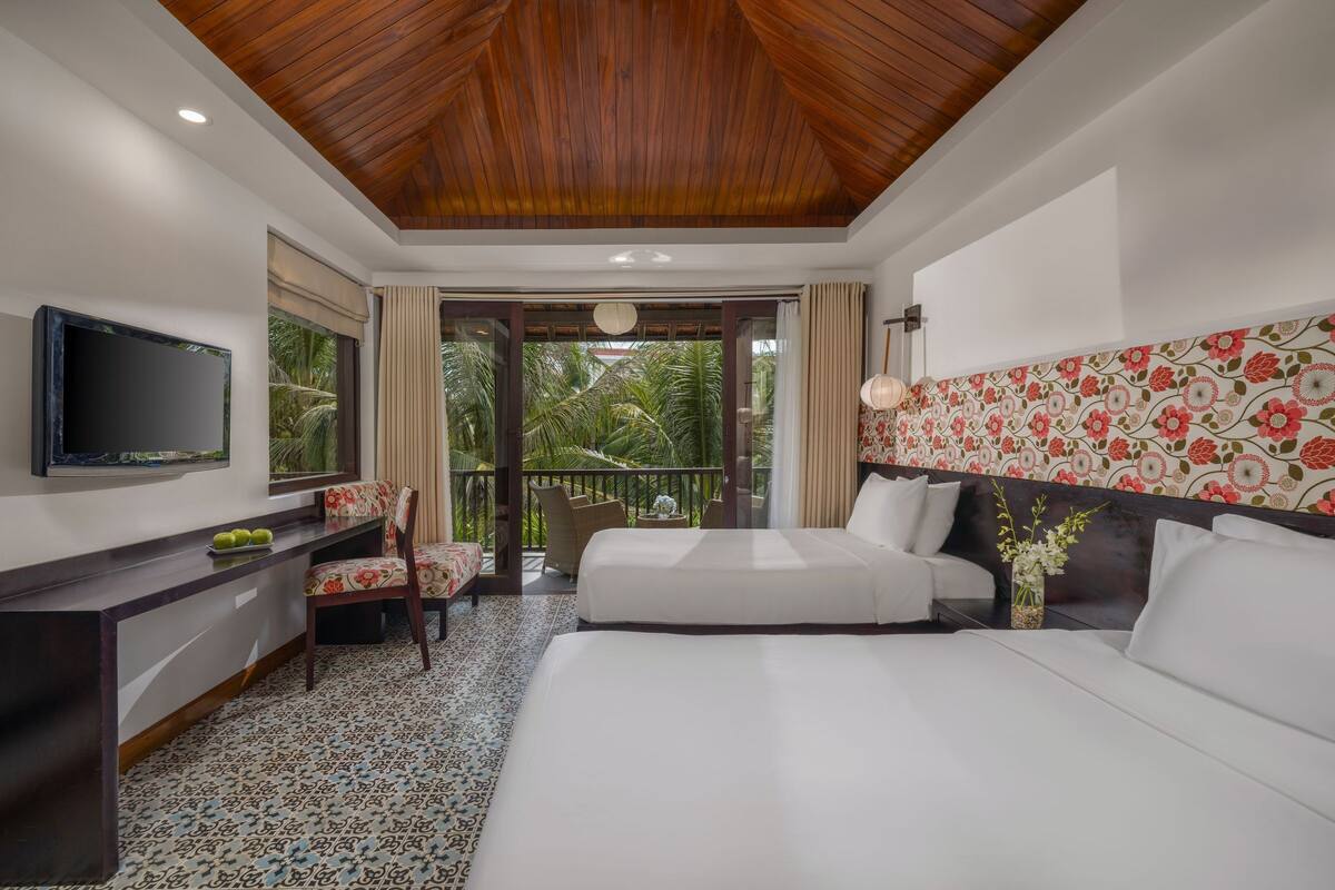 A hotel room with a wooden ceiling, a large bed, a desk with a television, and a balcony with a view of palm trees.