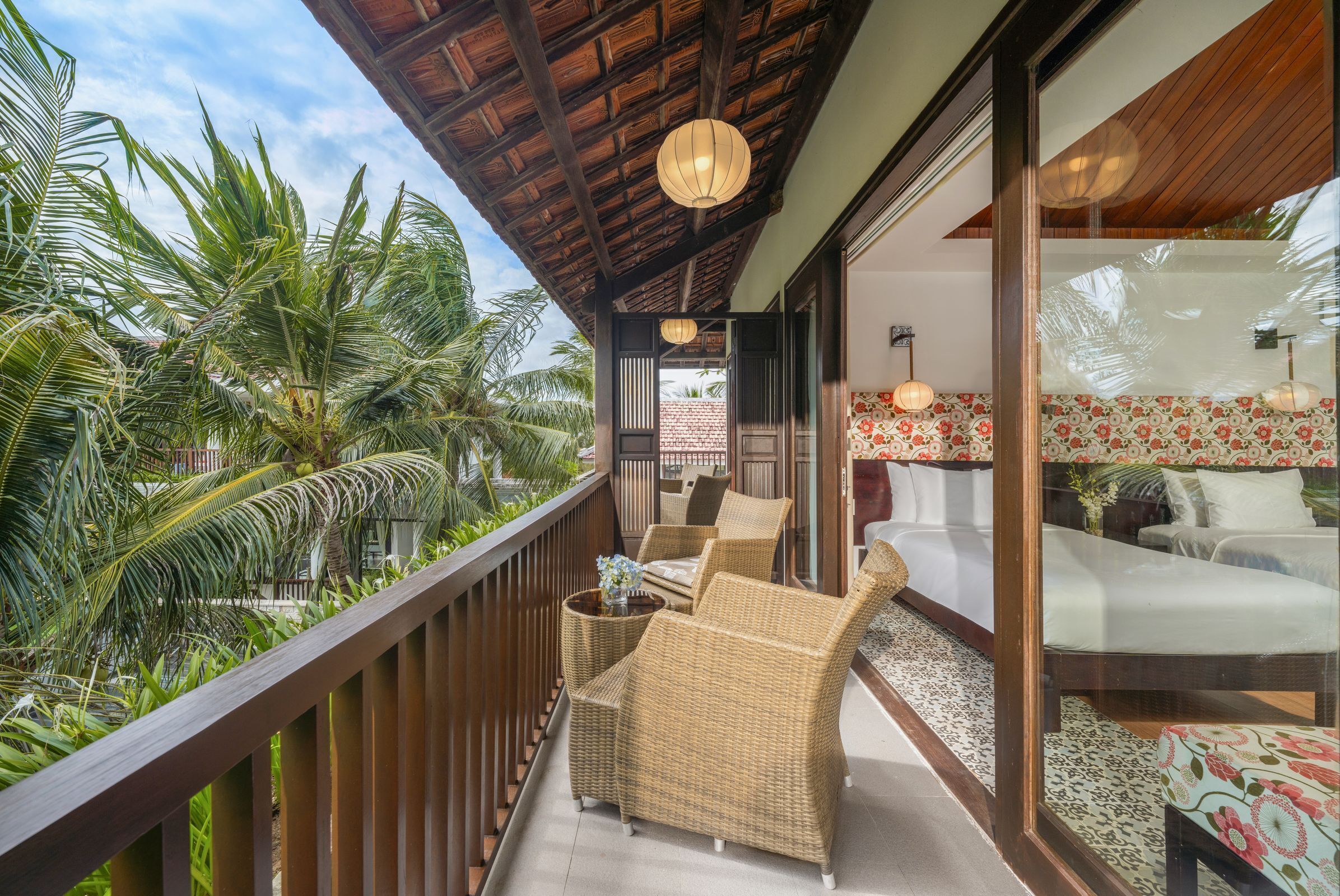 A balcony with wicker furniture, a view of palm trees, and a room with floral wallpaper and a bed.