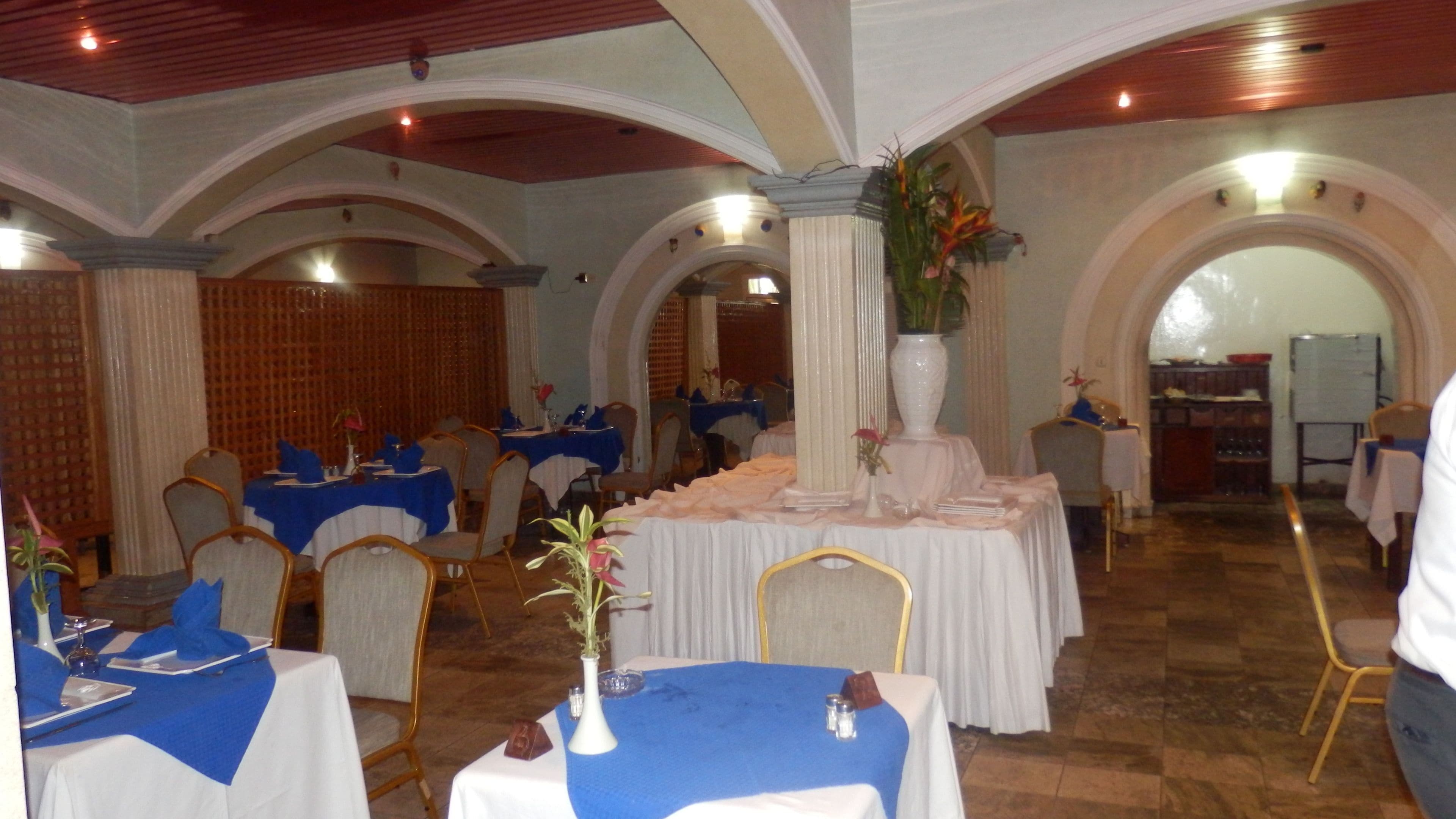 Een feestzaal met gedekte tafels, blauwe en witte tafelkleden, houten stoelen en een decoratieve pilaar in het midden.