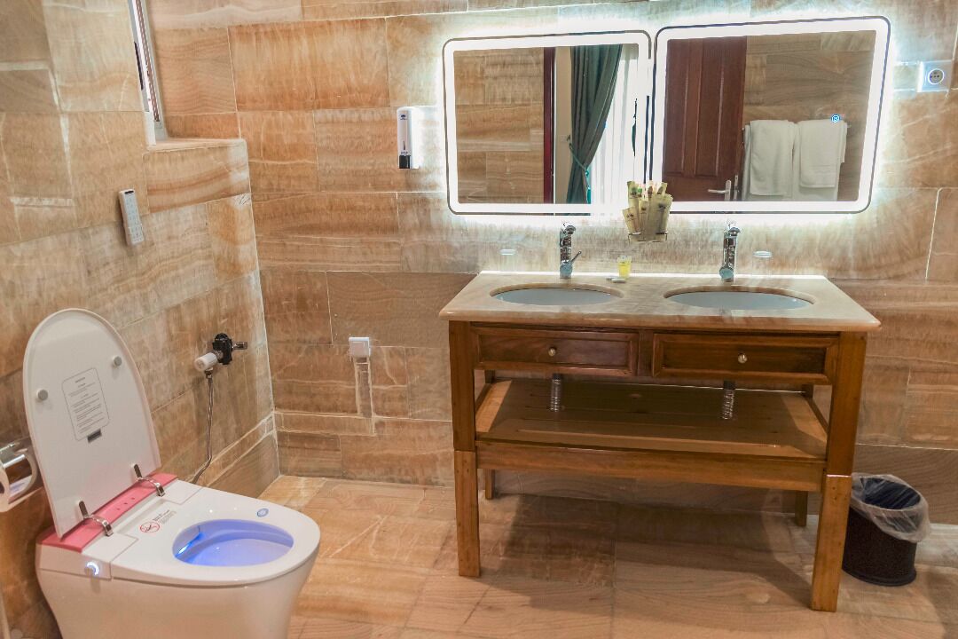 A bathroom with a modern design, featuring a wooden vanity with two sinks, a toilet with a blue-lit bowl, and a large mirror above the vanity.