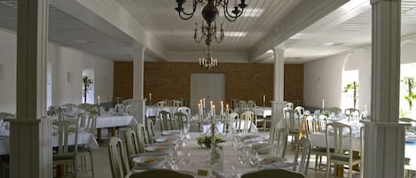 A spacious dining hall with white tables and chairs, a central chandelier, and brick walls.
